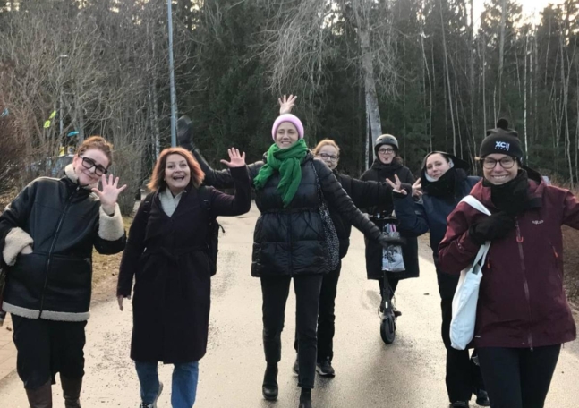 En grupp unga kvinnor går utomhus, ler och vinkar glatt mot kameran.