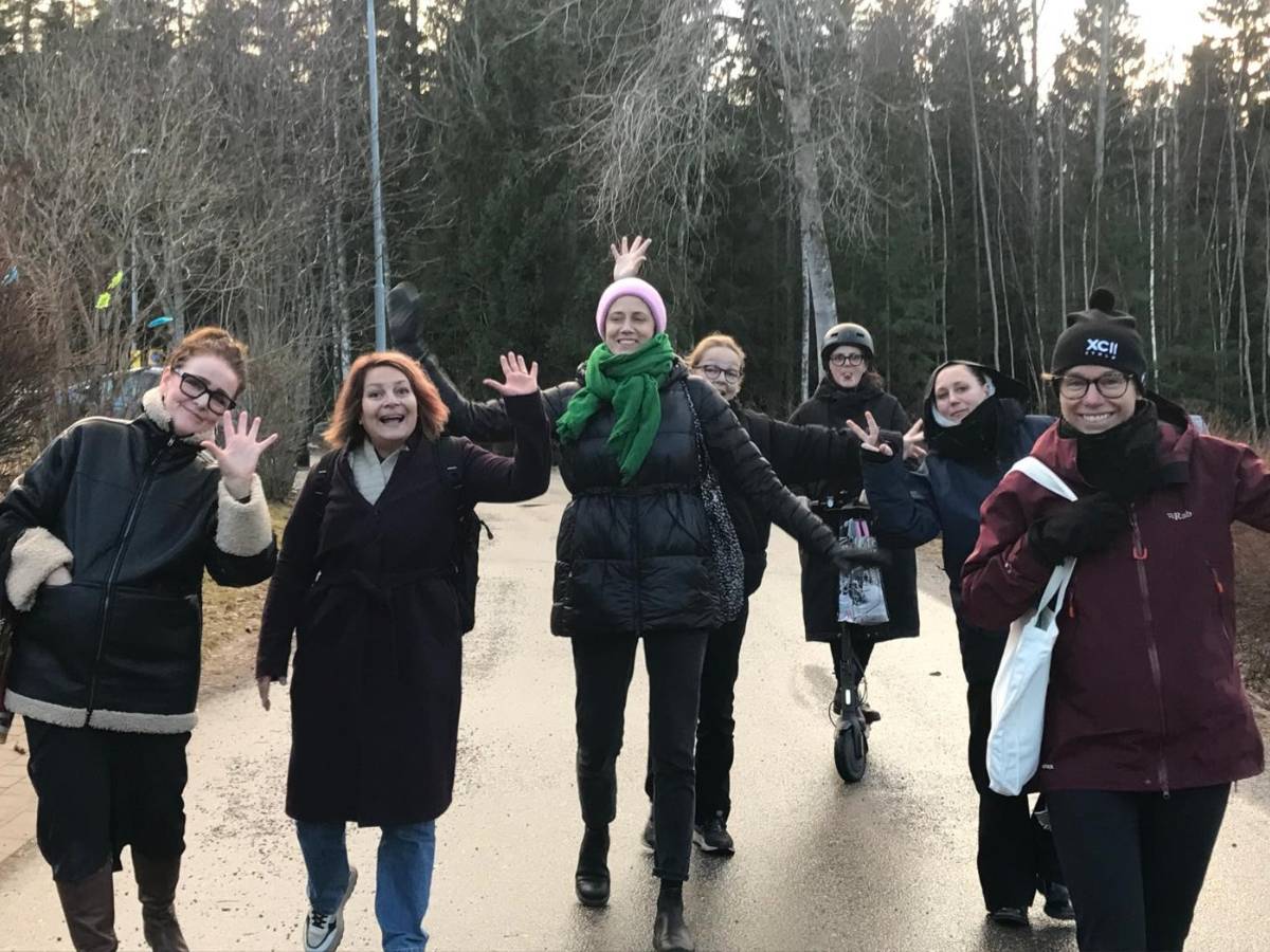 En grupp unga kvinnor går utomhus, ler och vinkar glatt mot kameran.