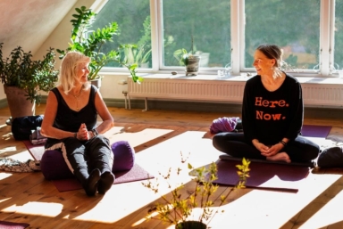 Två kvinnor sitter på varsin yogamatta i ett soligt rum med stora fönster.