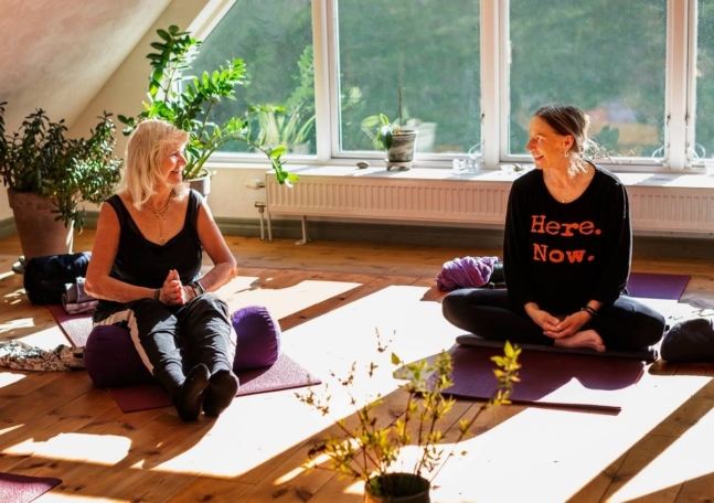 Två kvinnor sitter på varsin yogamatta i ett soligt rum med stora fönster.