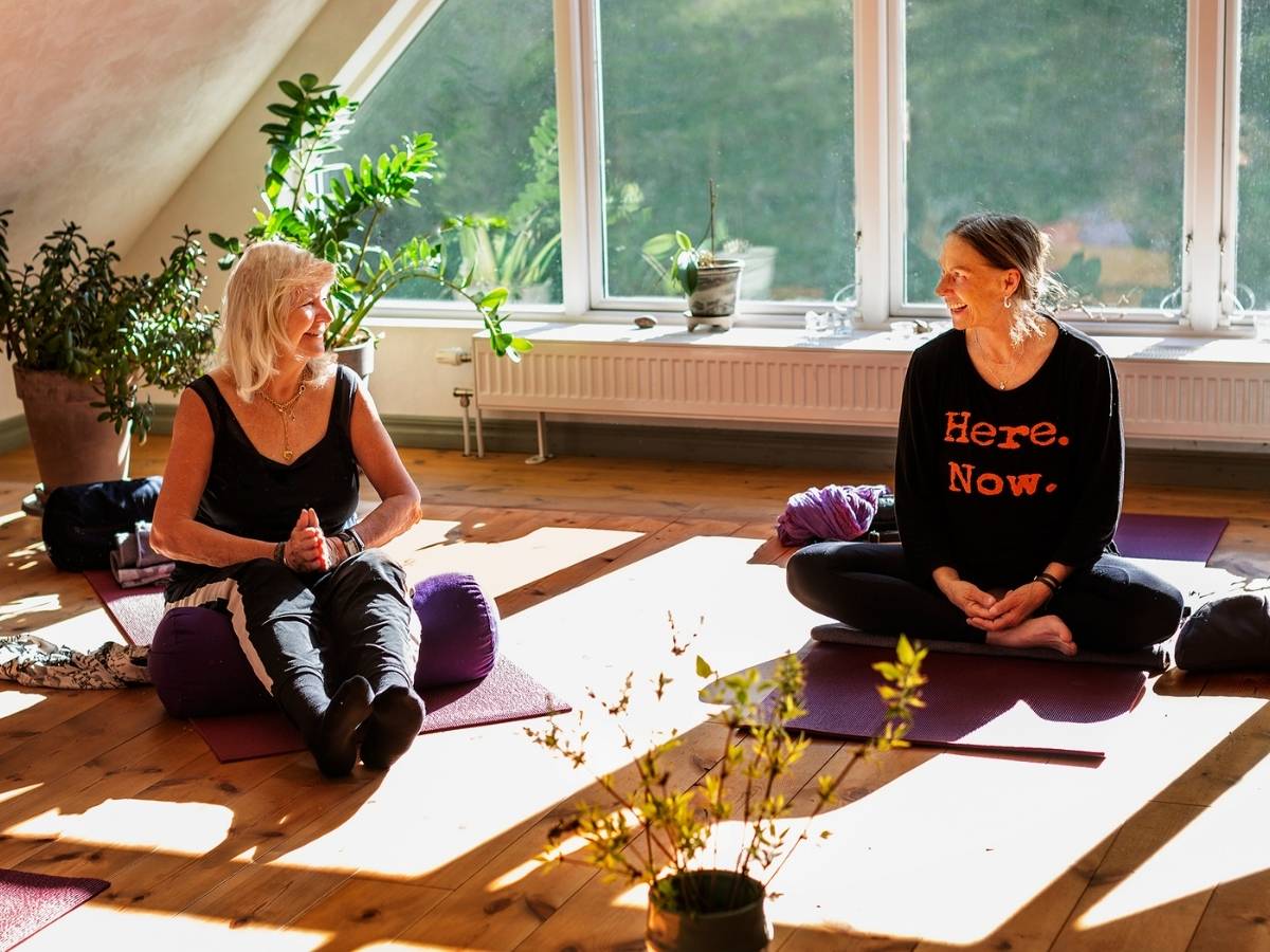 Två kvinnor sitter på varsin yogamatta i ett soligt rum med stora fönster.