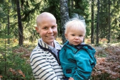 En kvinna utan hår står i skogen och håller i sitt barn.