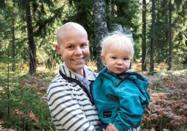 En kvinna utan hår står i skogen och håller i sitt barn.