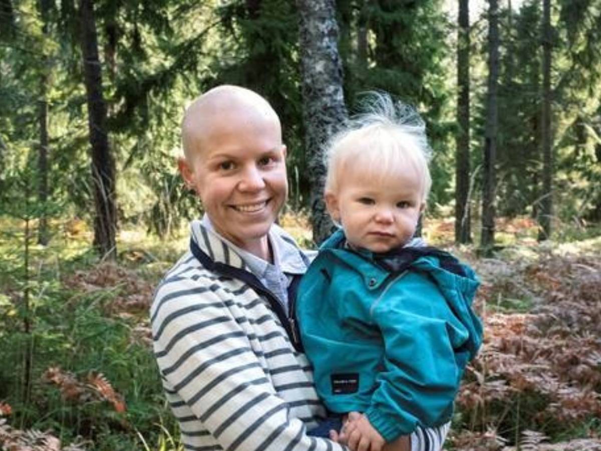 En kvinna utan hår står i skogen och håller i sitt barn.