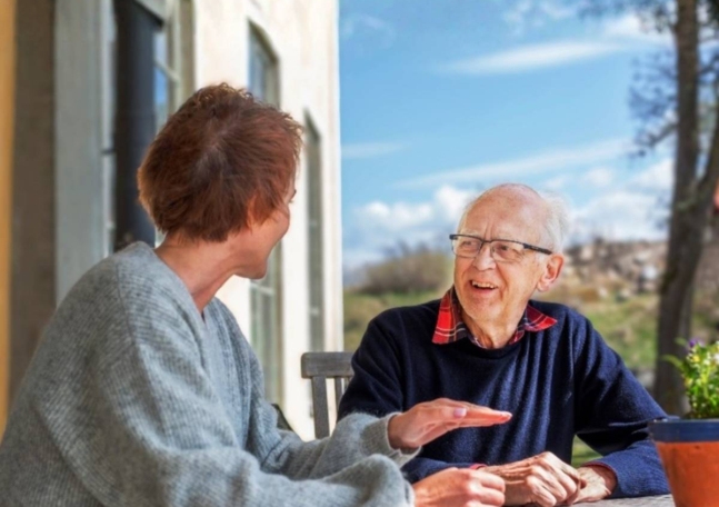 Ingemar Sjöberg sitter vid ett bord utomhus och samtalar med en deltagare på CancerRehabFondens rehabprogram.