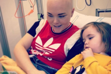 En kvinna utan hår sitter i en sjukhussäng och läser en bok för sin dotter.