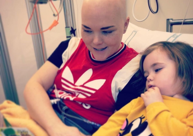 En kvinna utan hår sitter i en sjukhussäng och läser en bok för sin dotter.