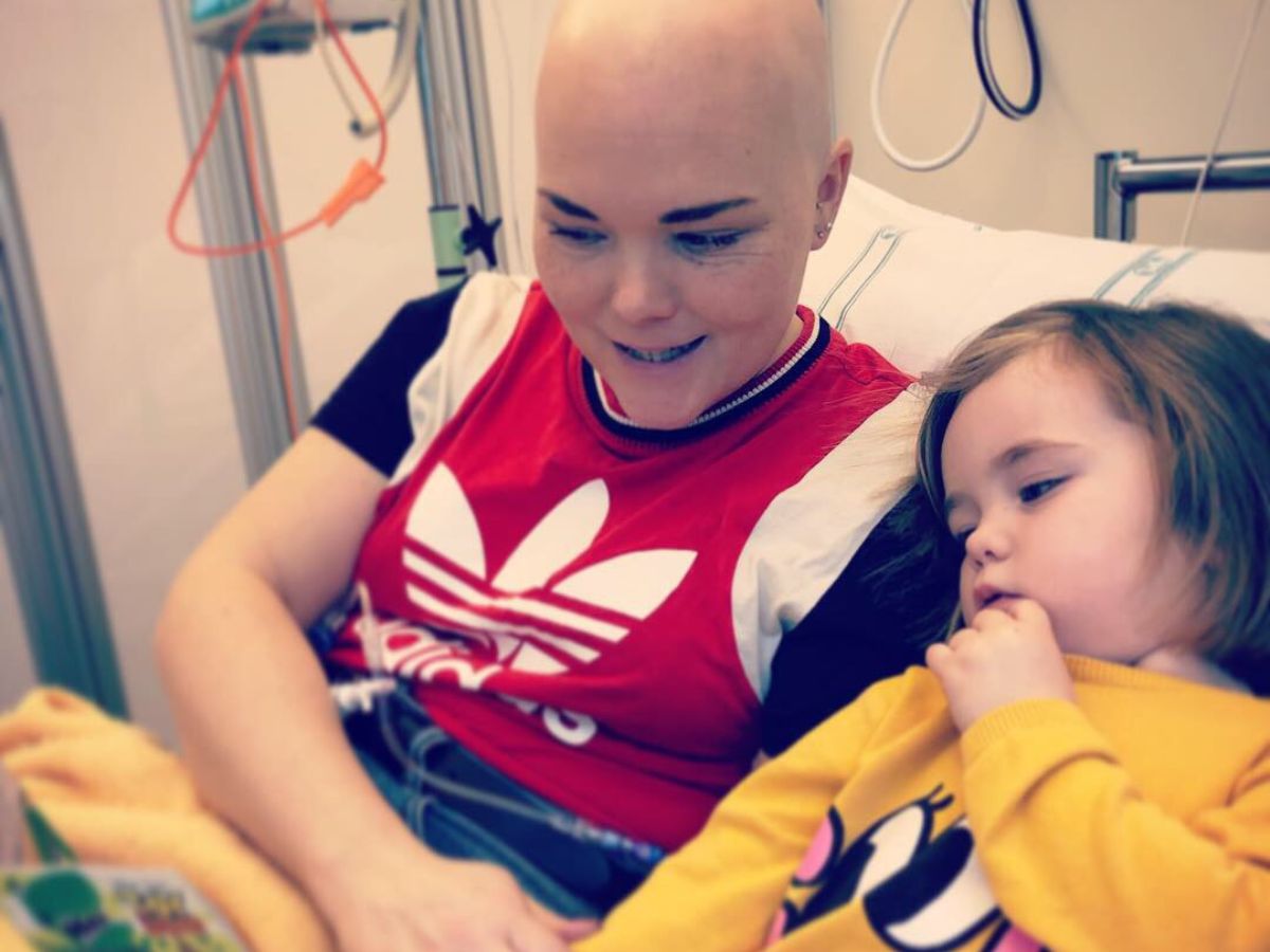 En kvinna utan hår sitter i en sjukhussäng och läser en bok för sin dotter.