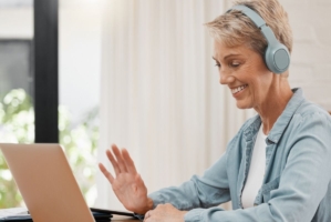 En kvinna med hörlurar sitter vid en laptop och vinkar till skärmen.