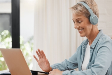 En kvinna med hörlurar sitter vid en laptop och vinkar till skärmen.