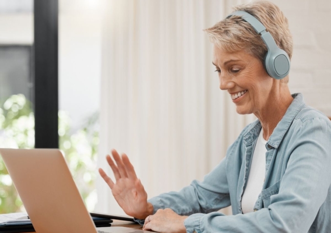 En kvinna med hörlurar sitter vid en laptop och vinkar till skärmen.