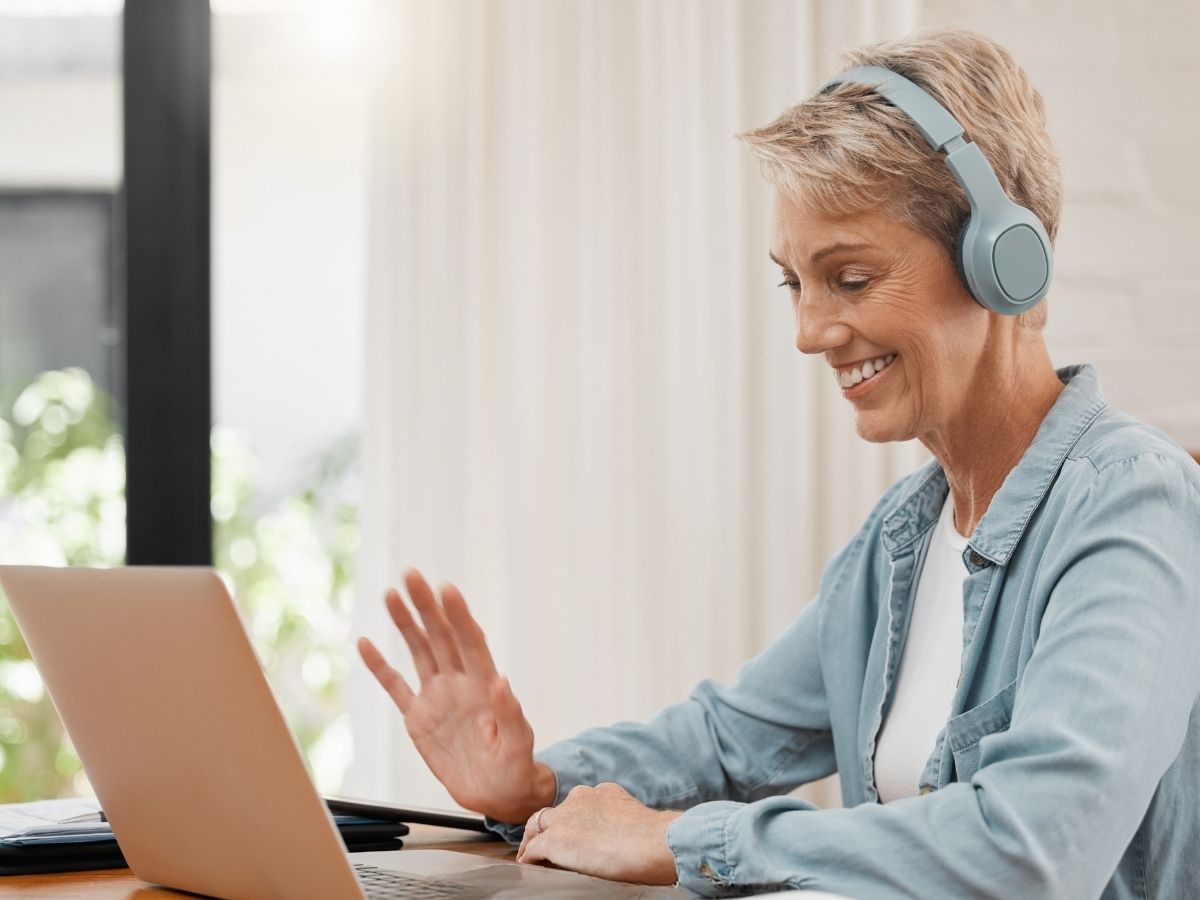 En kvinna med hörlurar sitter vid en laptop och vinkar till skärmen.