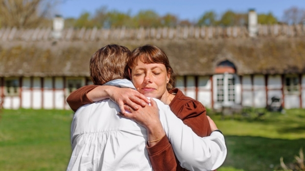 Två kvinnor kramas utomhus i en trädgård.