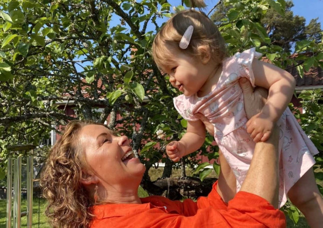 En kvinna ler och håller upp sitt barnbarn.