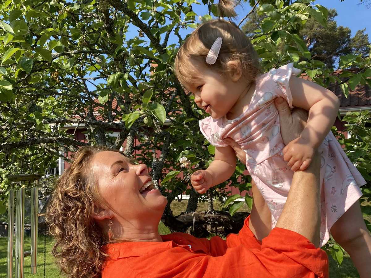 En kvinna ler och håller upp sitt barnbarn.